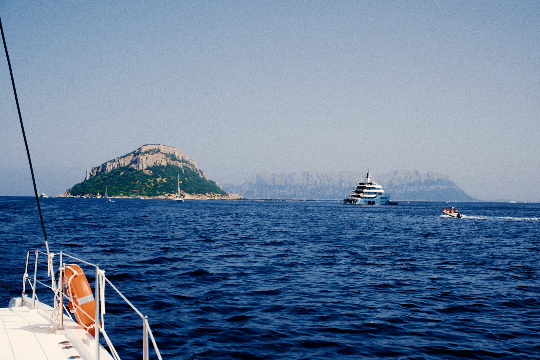A boat’s rail and life ring, the sea, an island and a far coast under a hazy sky. The whole frame on 35mm film at ISO 200.